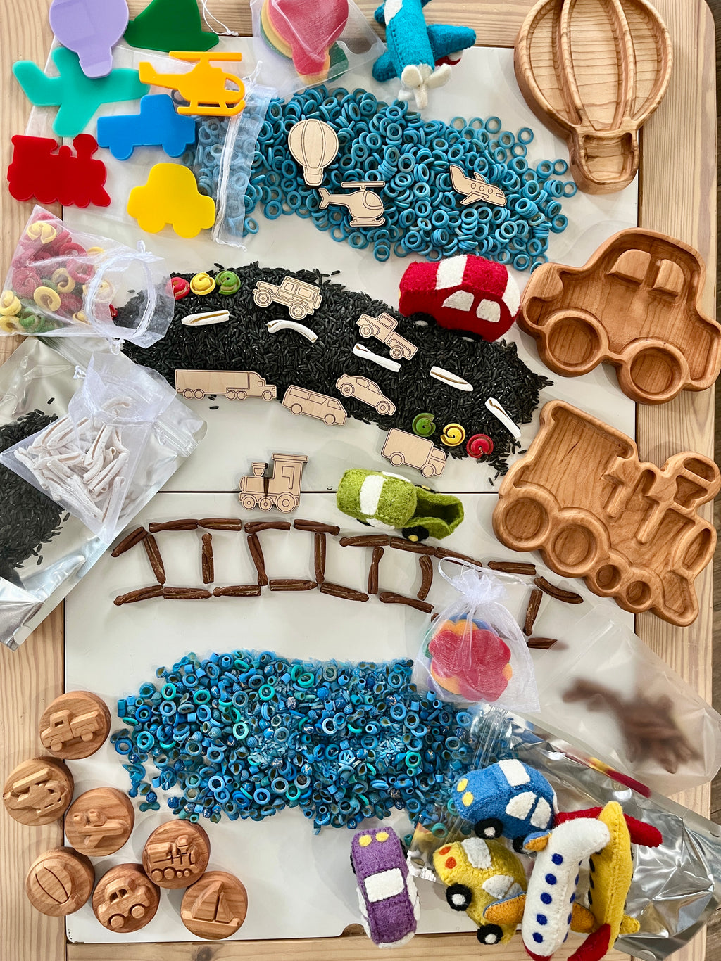 Wooden vehicle-shaped trays, colorful toy vehicles, and assorted sensory play materials arranged on a wooden surface.