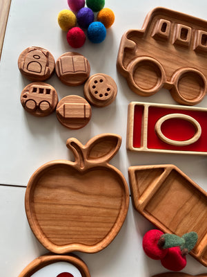 Set of wooden trays and tokens including apple-shaped tray, school bus tray, and felt balls in various colors on white surface