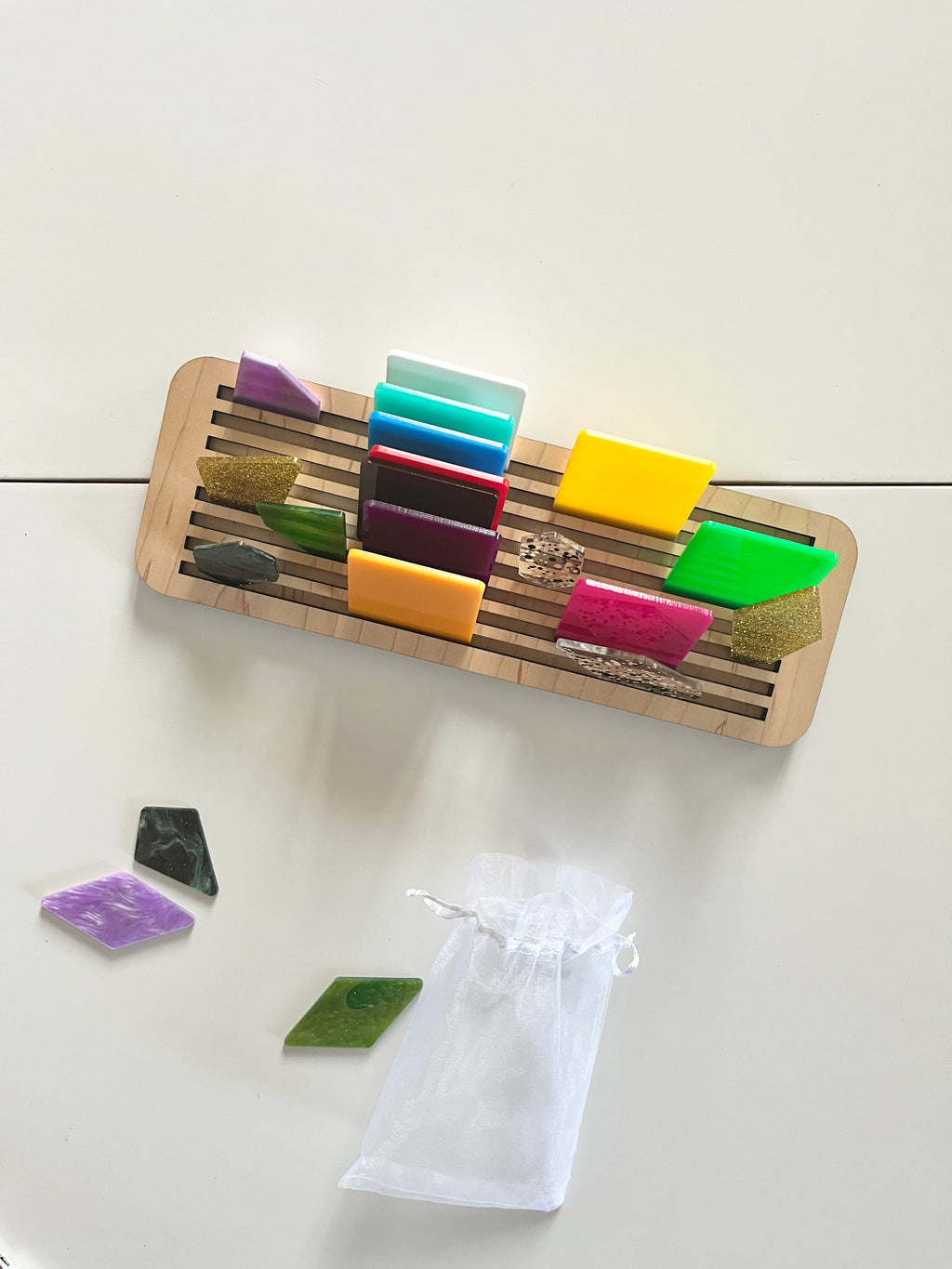 Wooden slotted rack holding colorful geometric acrylic pieces with several shapes and a white mesh pouch below on white surface