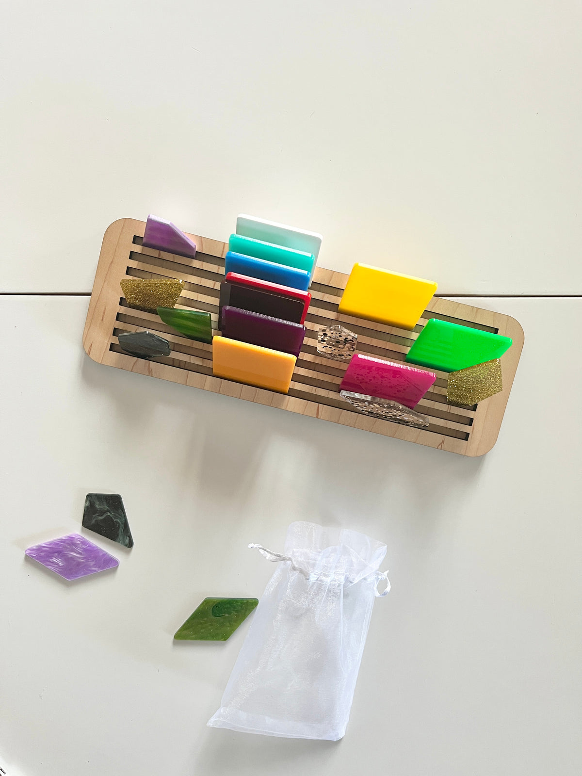 Wooden slotted rack holding colorful geometric acrylic pieces with several shapes and a white mesh pouch below on white surface