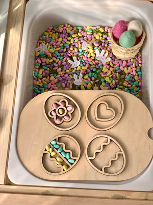Wooden tray with four egg-shaped cutouts over colorful craft beans, small bunny shapes, and a basket with pastel felt eggs