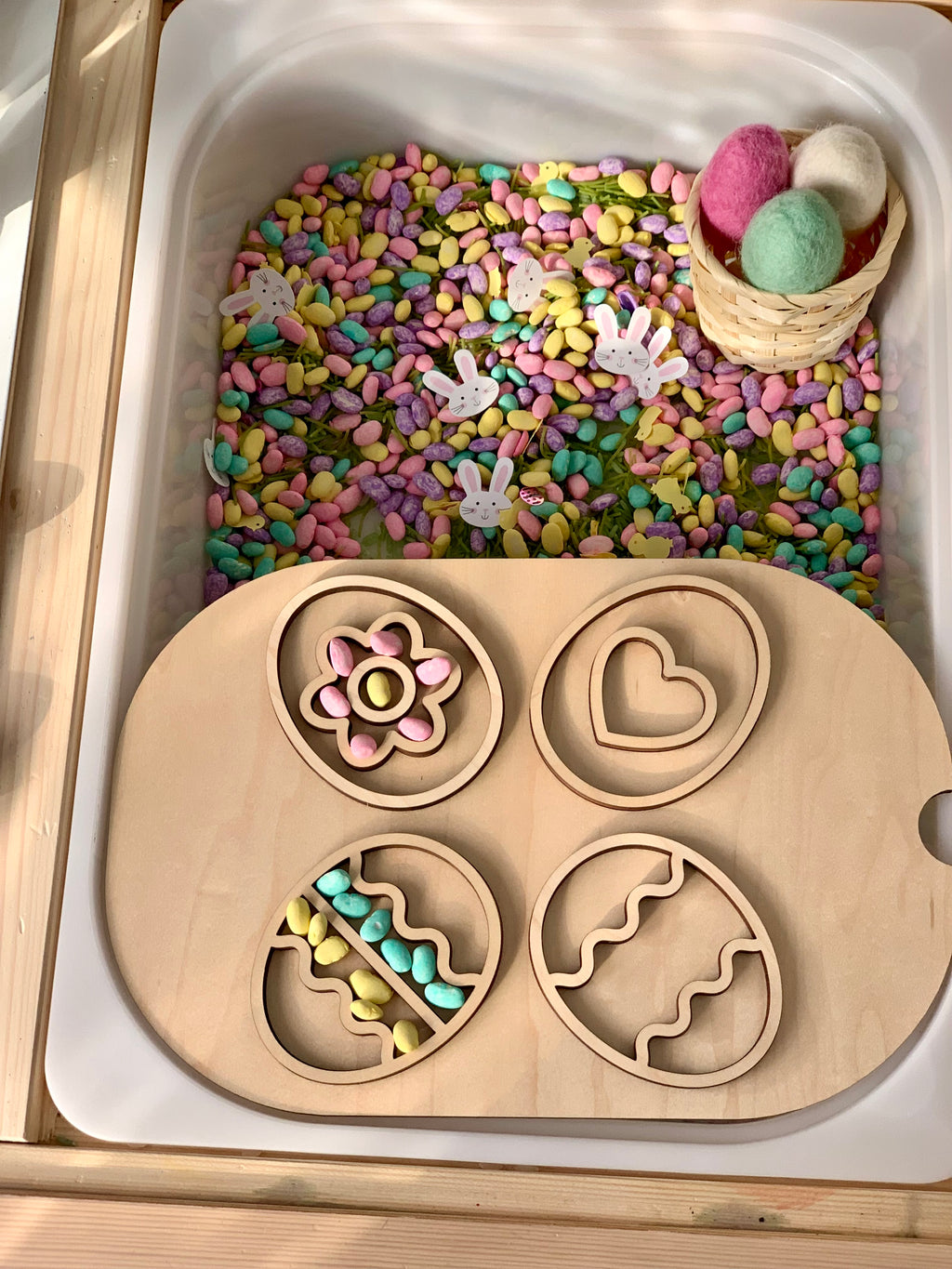 Wooden tray with four egg-shaped cutouts over colorful craft beans, small bunny shapes, and a basket with pastel felt eggs