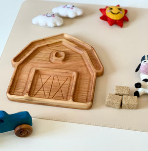 Wooden barn-shaped tray on beige surface surrounded by a blue toy tractor, felt cow, smiling sun and cloud decorations, and hay cubes