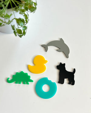 Five colorful acrylic shapes including gray dolphin, yellow duck, black dog, green dinosaur, and blue donut on white surface with green plant nearby