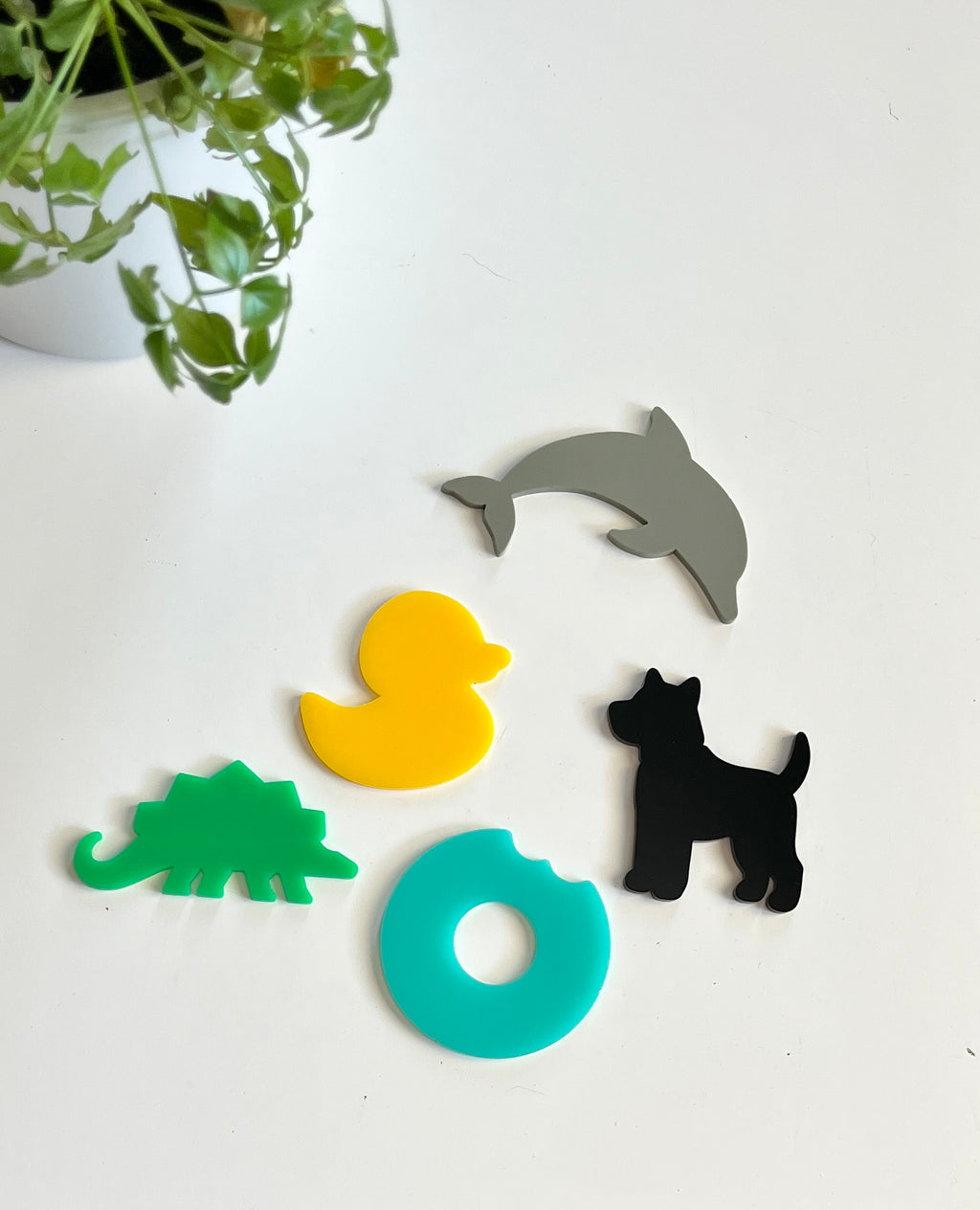 Five colorful acrylic shapes including gray dolphin, yellow duck, black dog, green dinosaur, and blue donut on white surface with green plant nearby