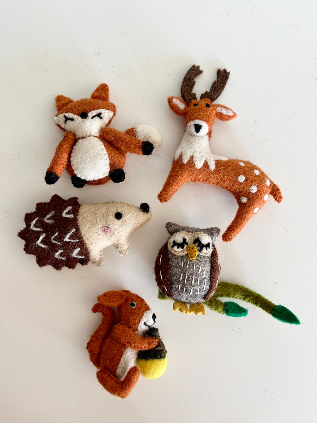 Five small felt animal figurines including a fox, deer, hedgehog, owl, and squirrel holding an acorn on a white surface