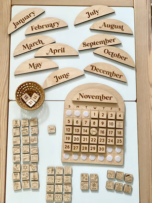 Wooden perpetual calendar set with month name plaques, numbered date tiles, and illustrated icon tiles arranged on white surface