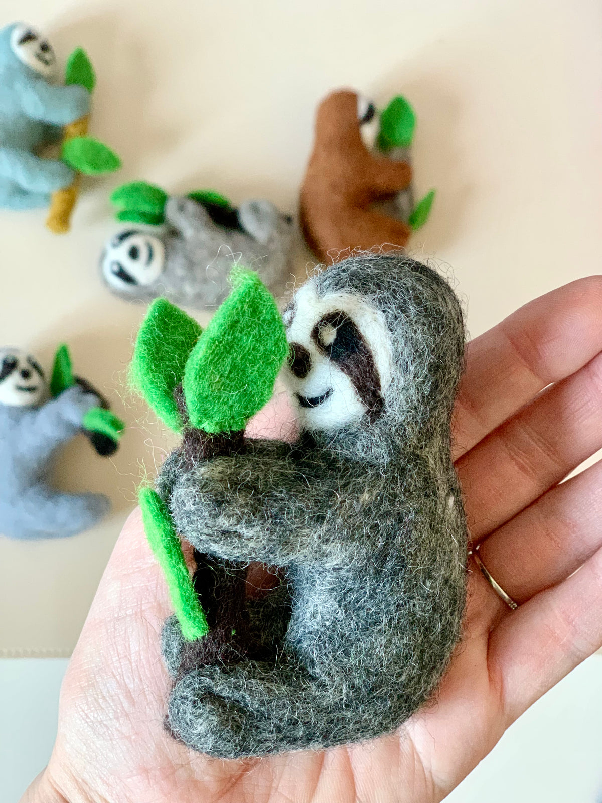 Hand holding a gray felt sloth figurine hugging a branch with green felt leaves against scattered sloth toys background