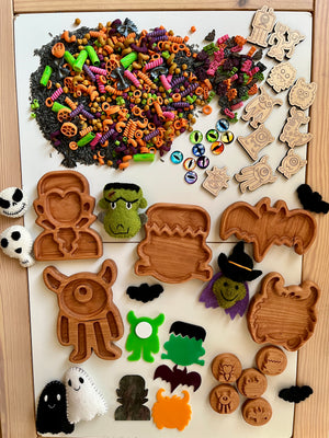 Halloween-themed wooden trays, tokens, colorful pasta shapes, monster eyes, and felt monster figures on white board and wooden table