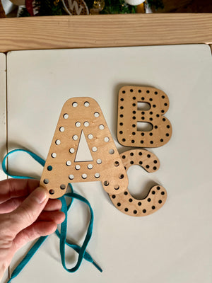 Hand holding a wooden letter A with holes and a blue lace for threading, alongside letters B and C, handcrafted homeschool education resources named Alphabet Lacing Letters (A, B, C shown — full alphabet optional).