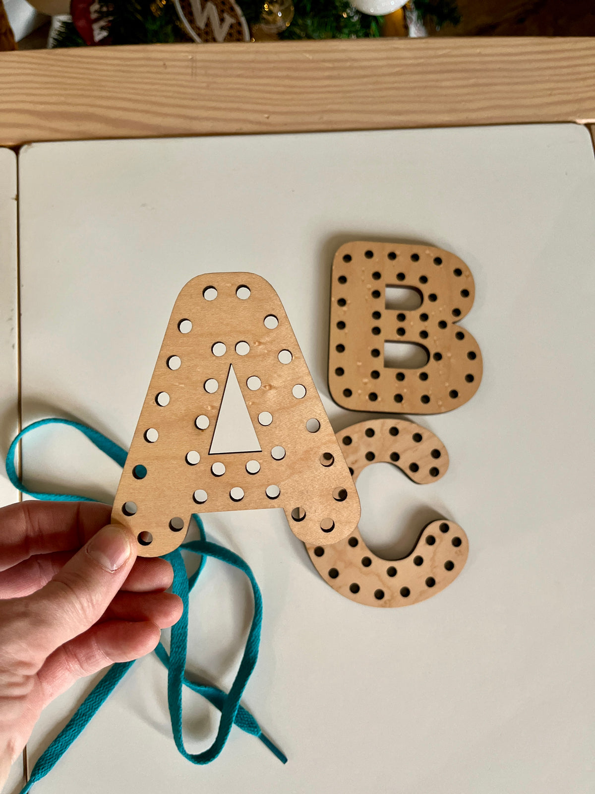 Hand holding a wooden letter A with holes and a blue lace for threading, alongside letters B and C, handcrafted homeschool education resources named Alphabet Lacing Letters (A, B, C shown — full alphabet optional).