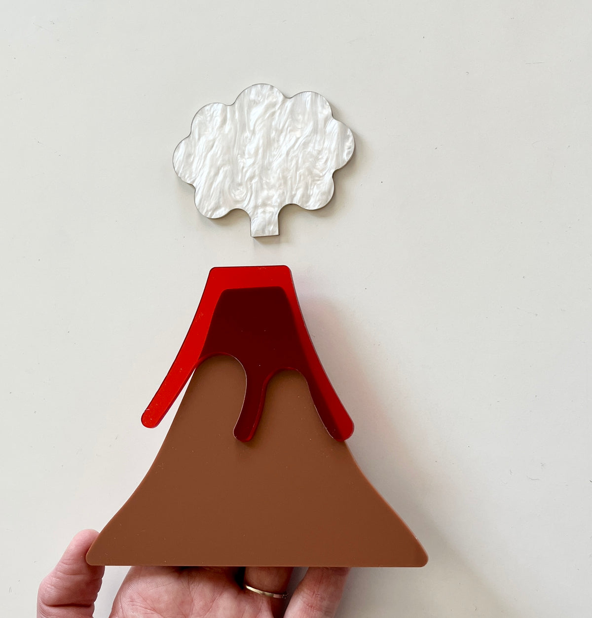 Hand holding layered acrylic volcano shape with brown base, red lava, and white cloud on white background