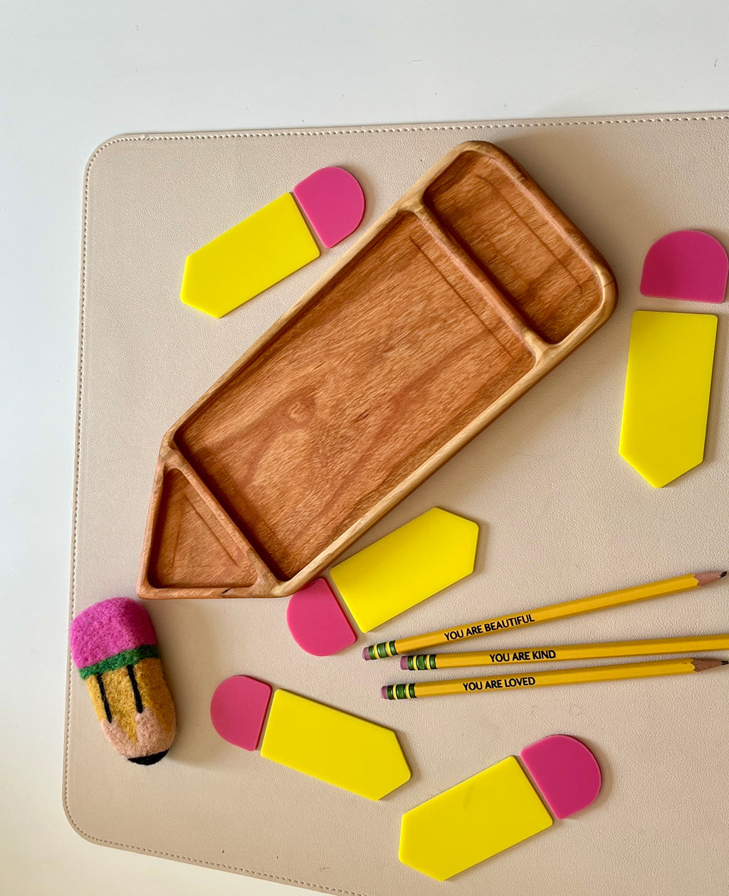 Carved wooden pencil-shaped tray surrounded by yellow and pink geometric pieces and three yellow pencils with positive messages on beige surface