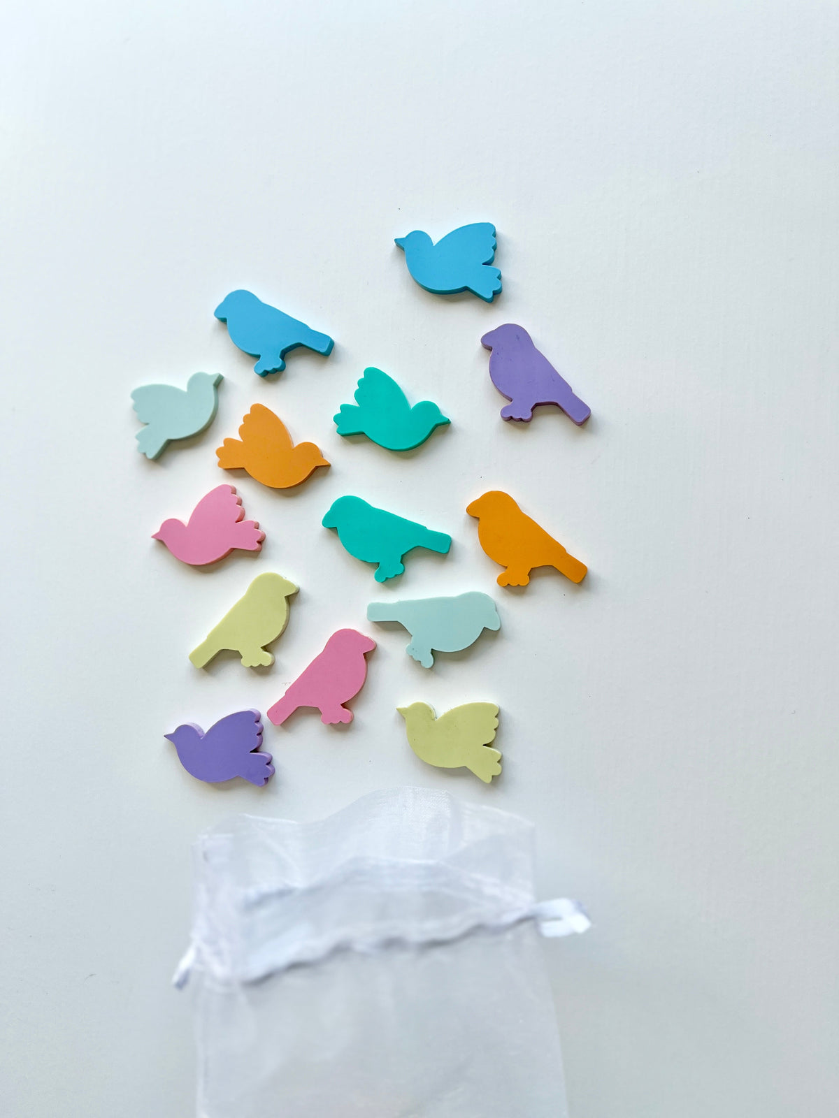 Colorful wooden bird-shaped game pieces scattered above a sheer white drawstring bag on a white background