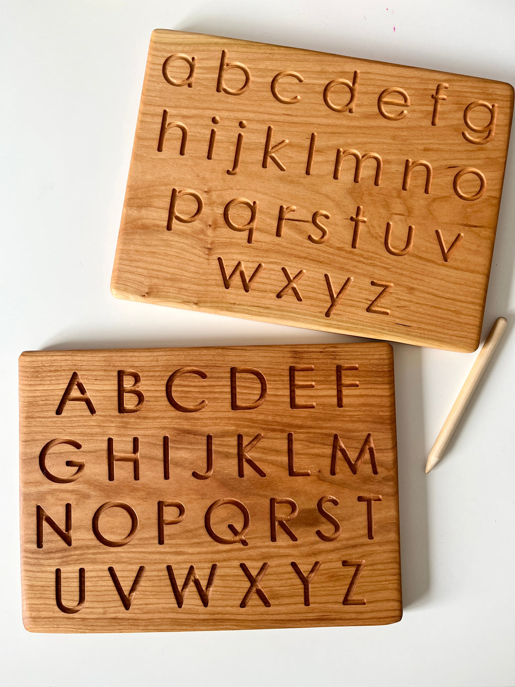 Two wooden tracing boards with carved uppercase and lowercase alphabet letters and a wooden stylus on white surface