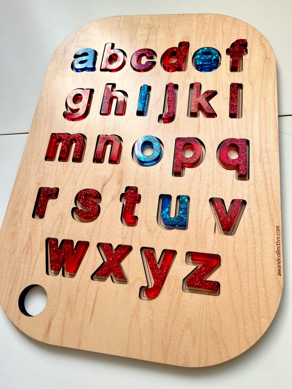 Lowercase resin alphabet letters in red and blue with glitter, set in a wooden puzzle board with rounded corners and a hole