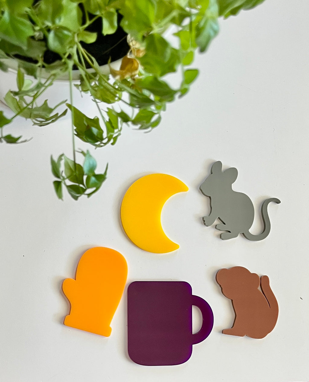 Acrylic shapes of yellow crescent moon, gray mouse, orange mitten, purple mug, and brown dog on white surface with green plant above