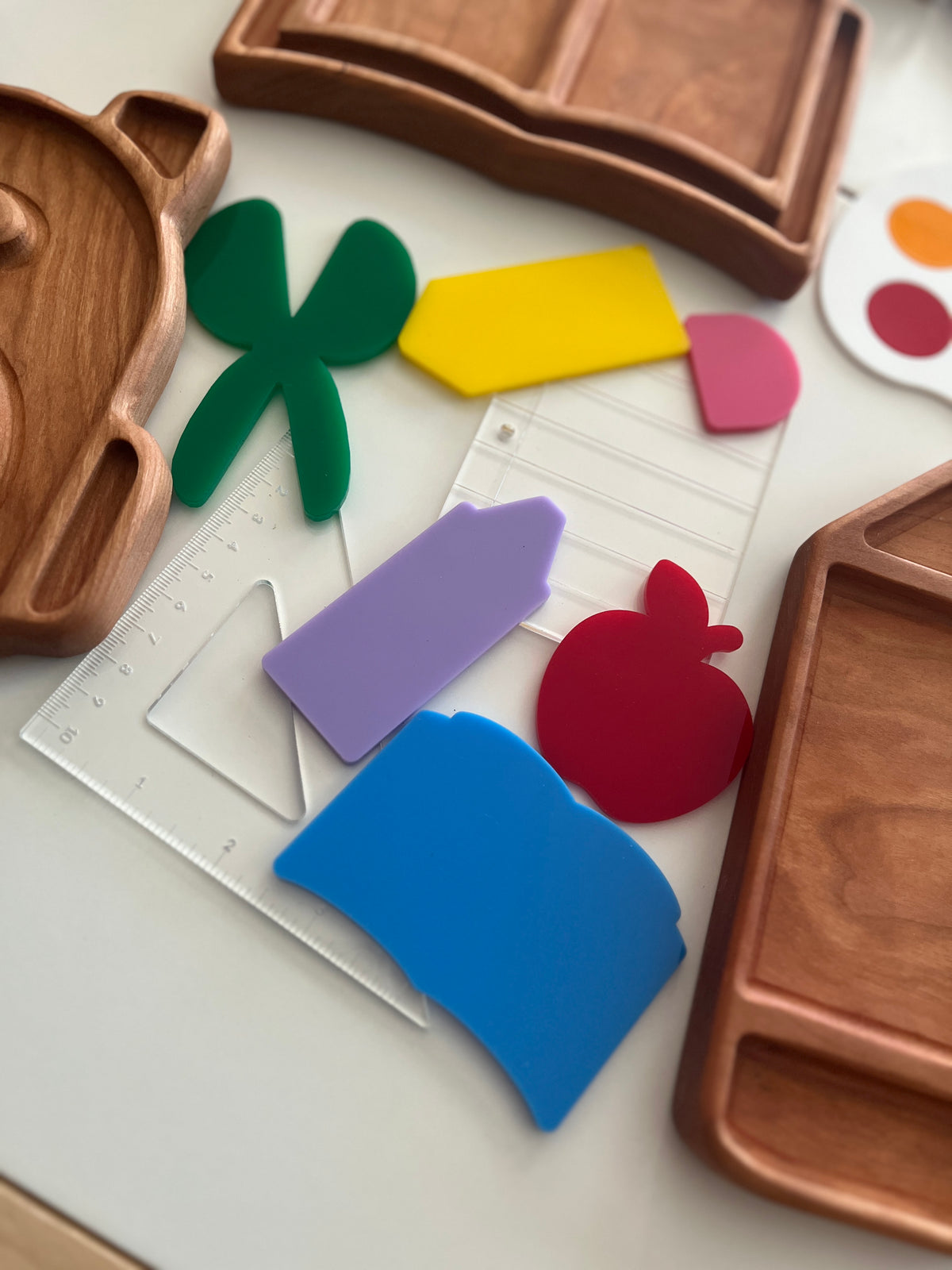 Colorful acrylic shapes including apple, bow, hat, and rectangles arranged with wooden trays and clear ruler on white surface