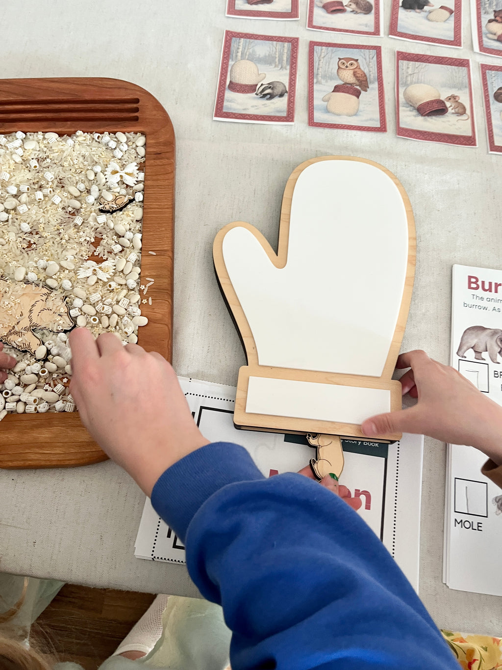 The Mitten Wooden Animals and/or Little Dry Erasables with Mitten Interactive Learning Set showing a child’s hands engaging with a wooden mitten-shaped dry-erase board, wooden tray with sensory filler, and illustrated animal cards, handcrafted homeschool educational resources.