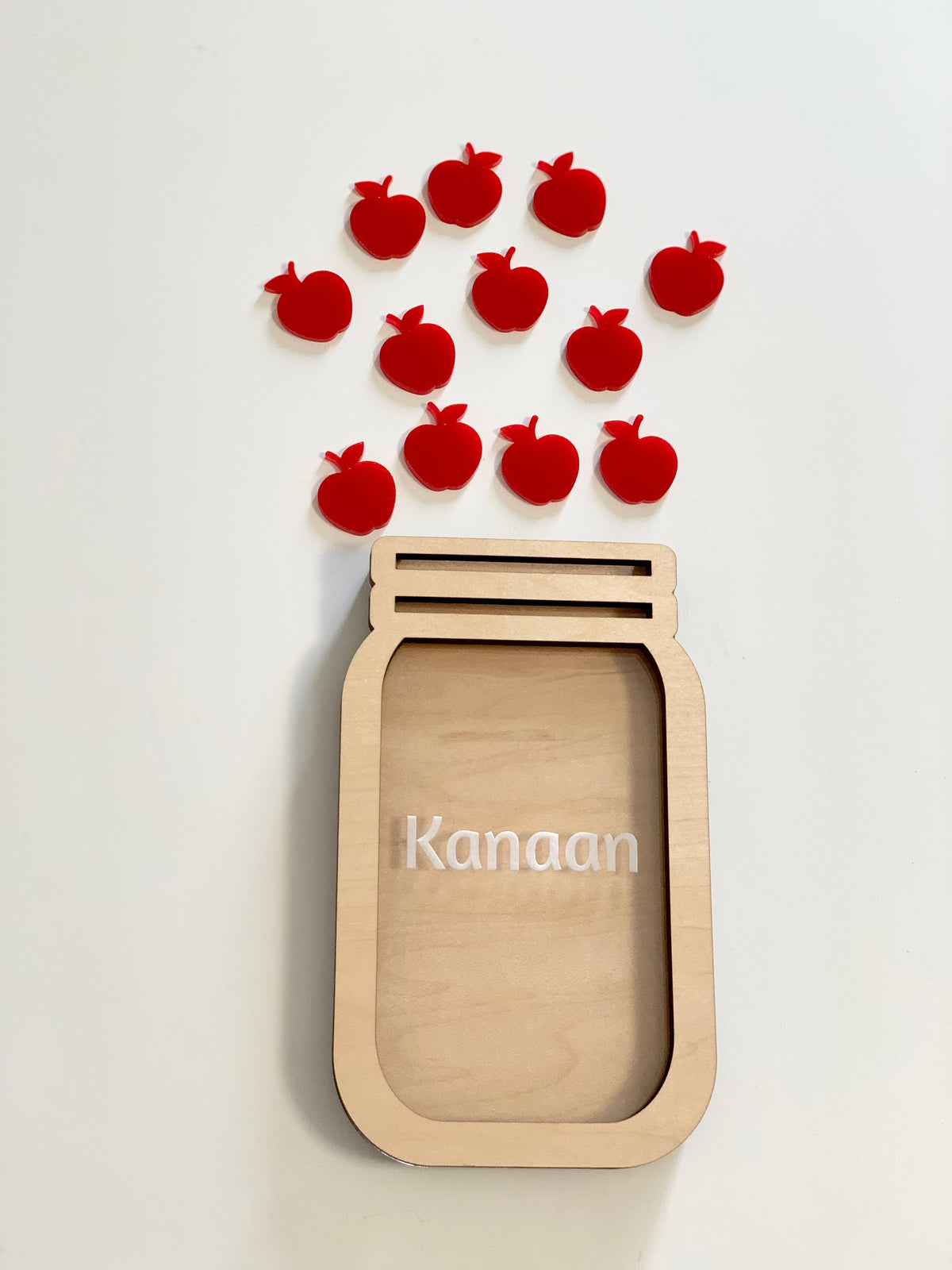 Wooden jar-shaped frame with the name Kanaan and scattered red apple-shaped tokens above on white background