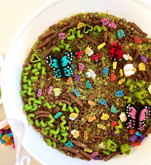 White bowl filled with colorful felt insects, wooden pasta, and green spiral pasta scattered on dried grass mix