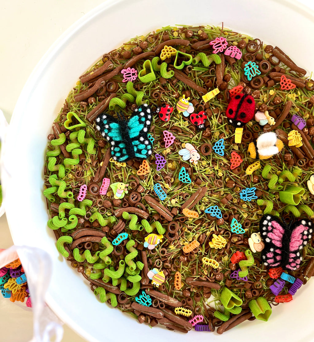 White bowl filled with colorful felt insects, wooden pasta, and green spiral pasta scattered on dried grass mix