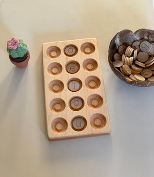 Light wooden tray with round indentations holding wooden letter pieces, next to a small crocheted cactus and coconut bowl with tokens