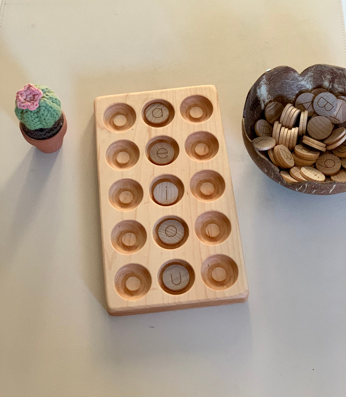Light wooden tray with round indentations holding wooden letter pieces, next to a small crocheted cactus and coconut bowl with tokens
