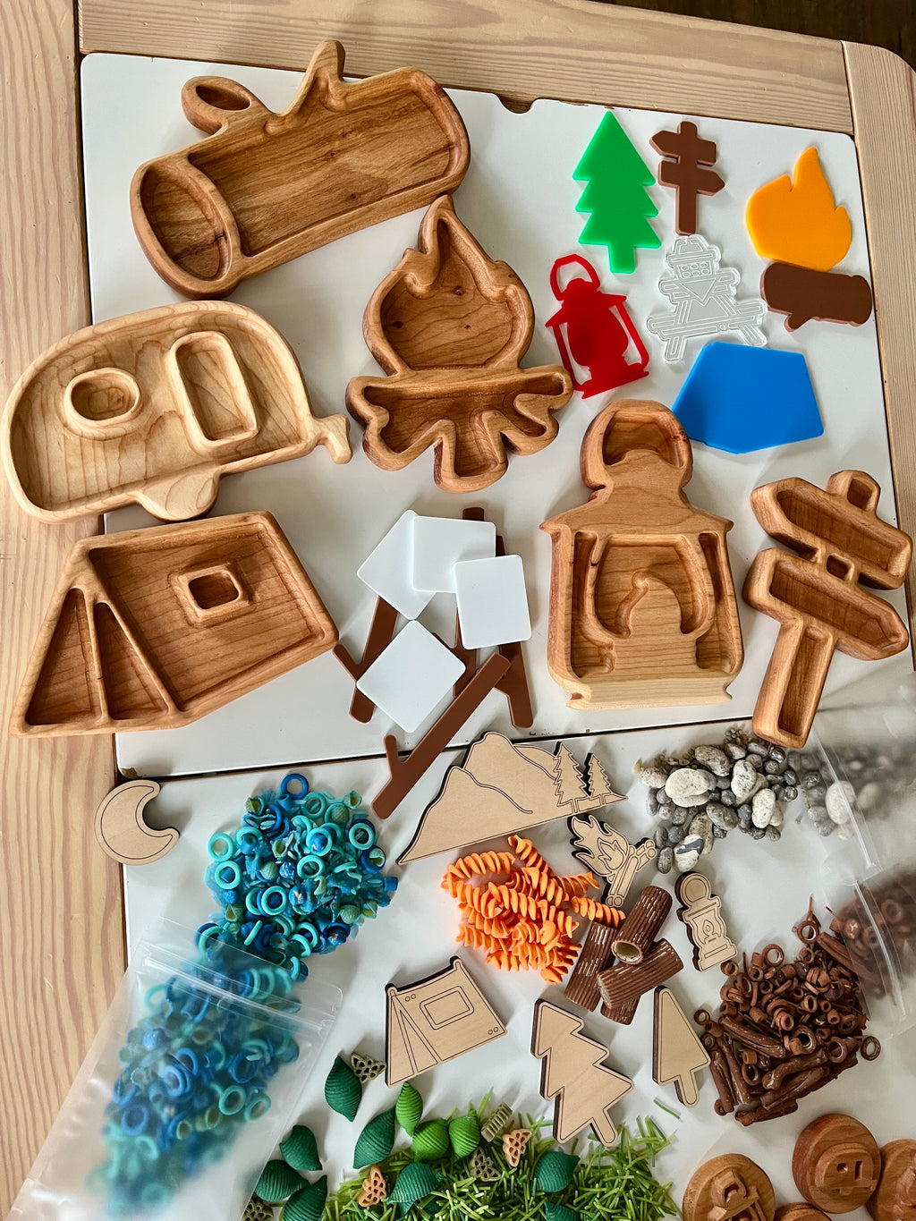 Wooden camping-themed trays shaped like trailer, campfire, lantern, tent, signpost surrounded by colorful nature play items on table