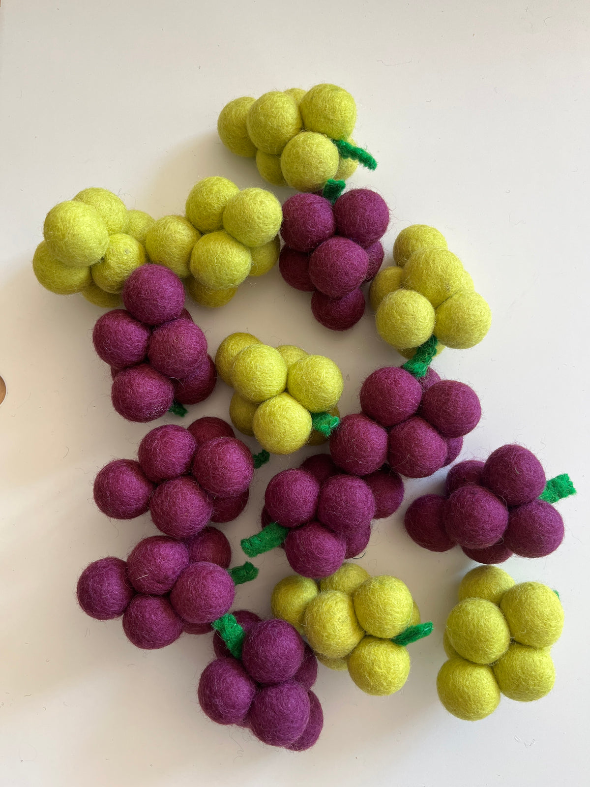 Clusters of small felted balls in yellow and purple arranged to resemble bunches of grapes on a white surface