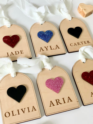 Set of wooden gift tags with glittery and solid heart embellishments and names engraved, tied with white fabric ribbons