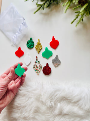 Hand holding green acrylic ornament cutout with various colorful glitter and solid acrylic ornament shapes nearby on white surface