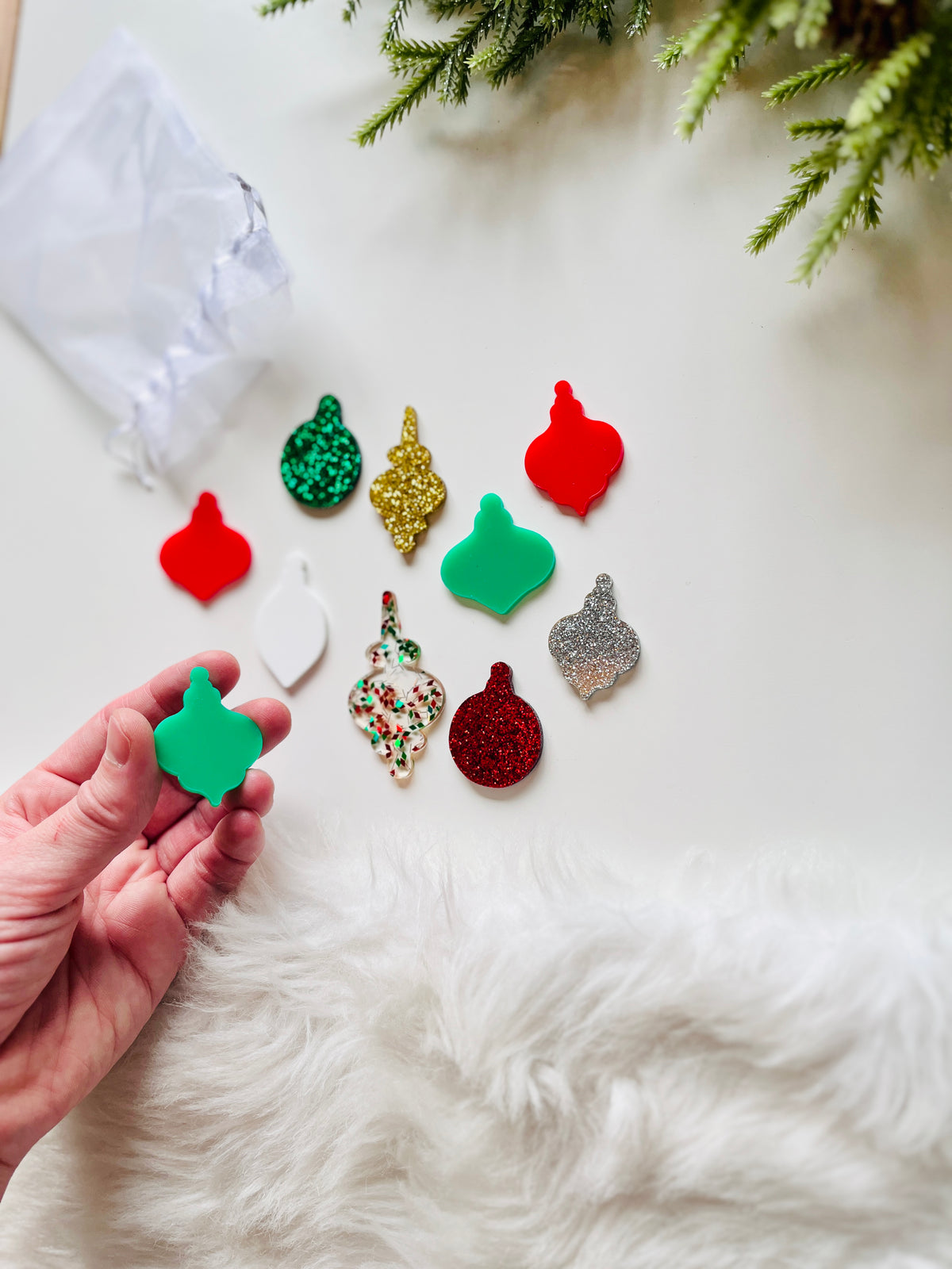 Hand holding green acrylic ornament cutout with various colorful glitter and solid acrylic ornament shapes nearby on white surface