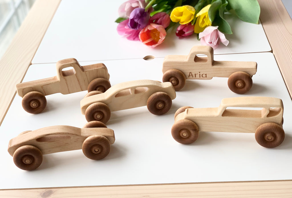Five handcrafted wooden toy vehicles arranged on a white surface with colorful tulips in the background