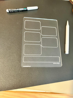 Transparent plastic writing board with rectangular sections placed on a black surface alongside two white markers