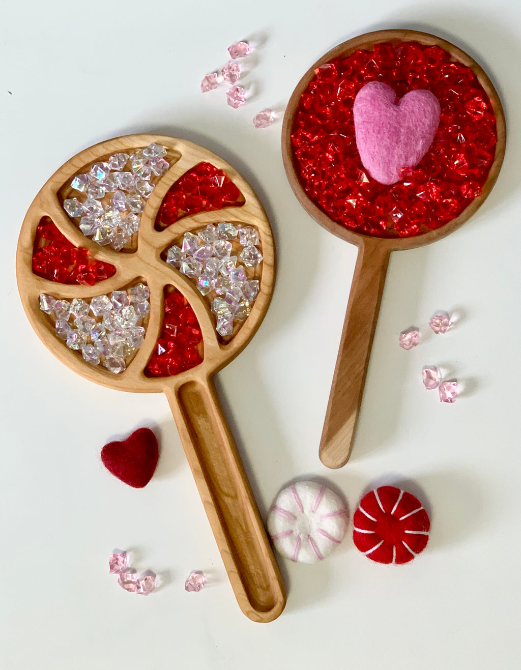 Two wooden paddle trays filled with red and clear acrylic gems and felt heart and peppermint shapes around them on white surface