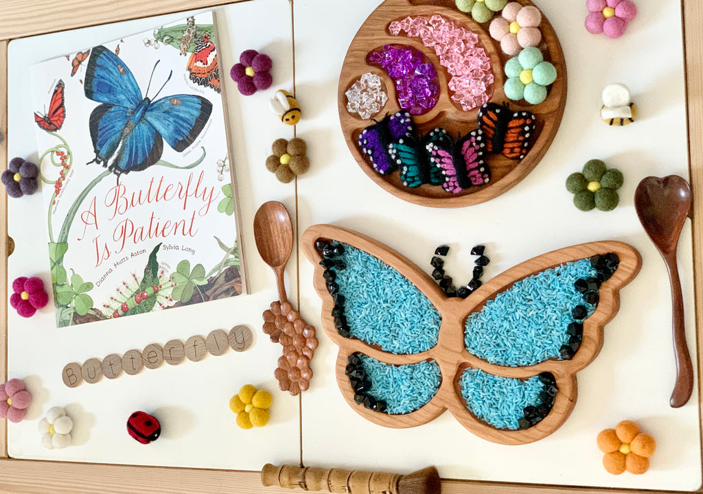 Wooden butterfly-shaped sensory tray filled with blue and black beads, surrounded by butterfly toys, flowers, and a butterfly book on white table