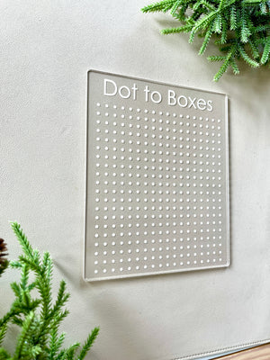 Clear acrylic Dot to Boxes game board with engraved grid dots on beige surface surrounded by green foliage