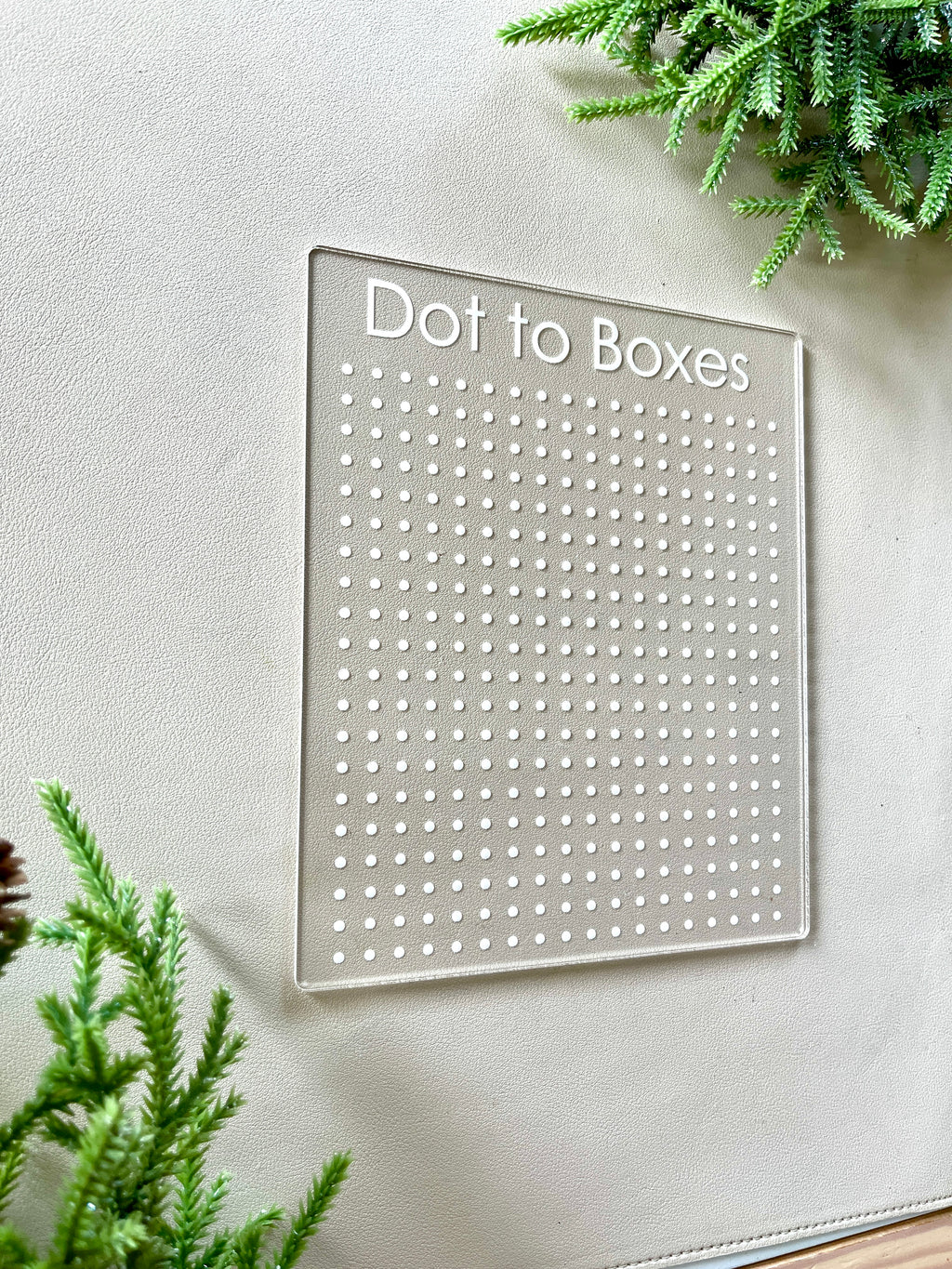Clear acrylic Dot to Boxes game board with engraved grid dots on beige surface surrounded by green foliage