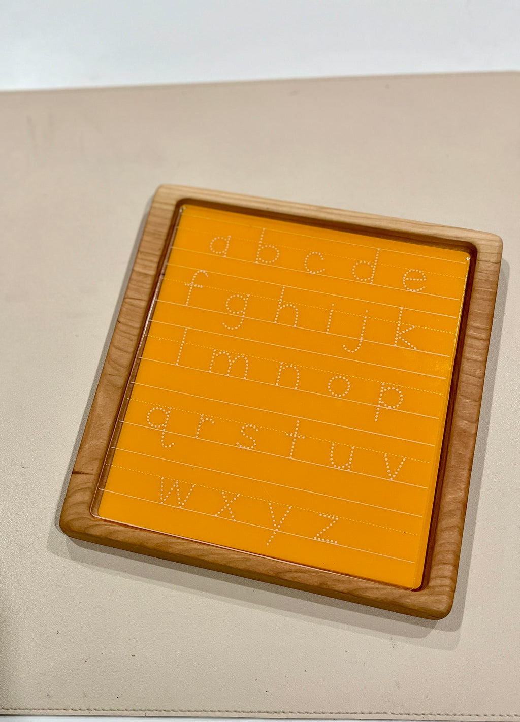 Wooden-framed orange acrylic board with dotted lowercase alphabet letters for tracing practice on beige surface