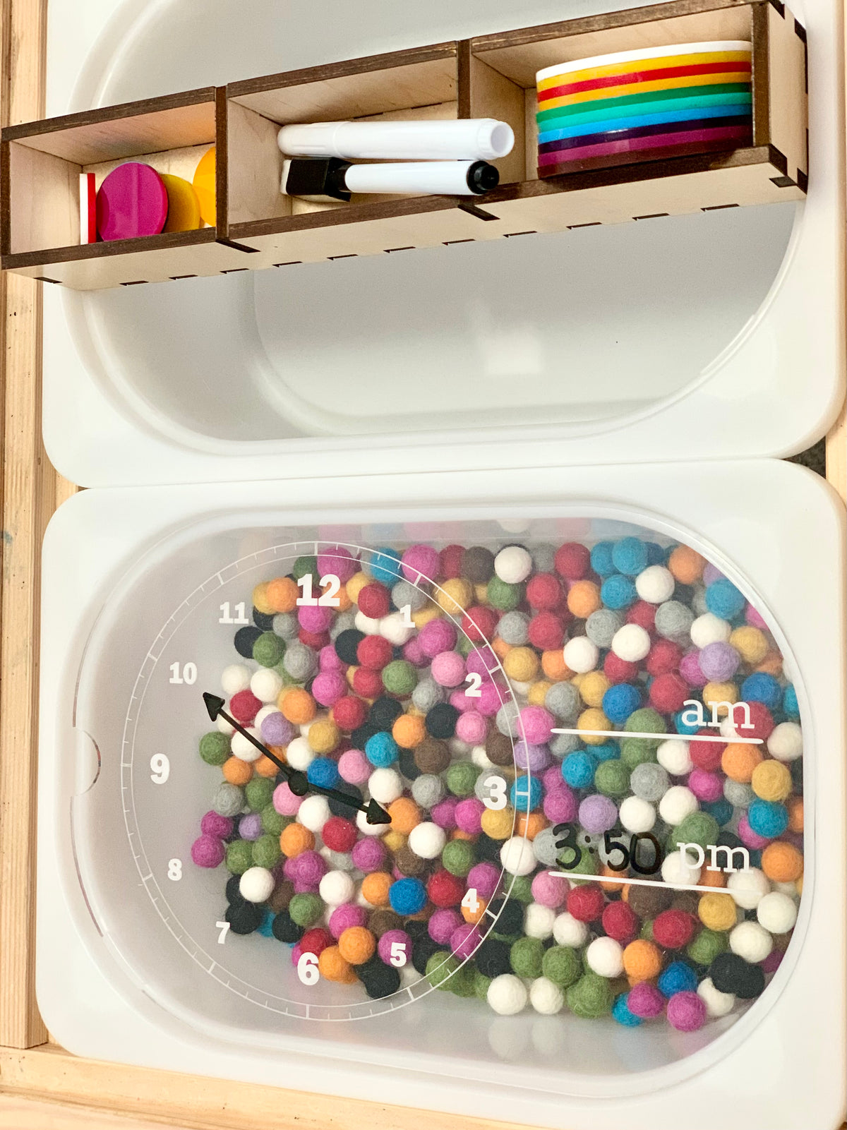 Clear plastic learning clock with colorful felt balls inside and dry erase markers stored above in wooden compartments