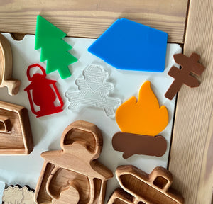 Colorful plastic camping shapes including tree, tent, lantern, campfire, signpost, and wooden stamp outlines on light surface