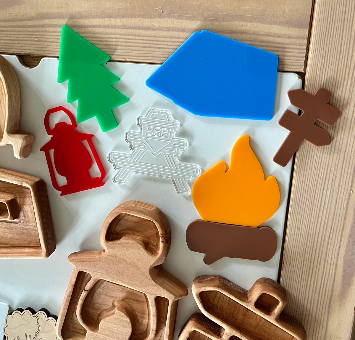 Colorful plastic camping shapes including tree, tent, lantern, campfire, signpost, and wooden stamp outlines on light surface