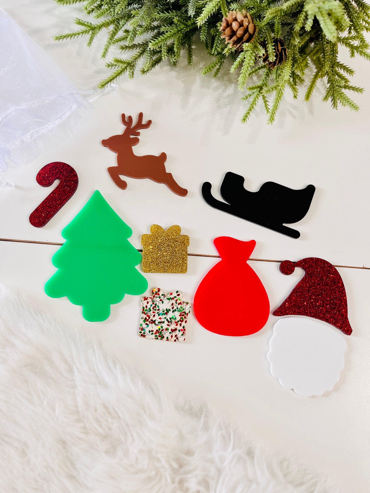 Assorted flat Christmas-themed acrylic shapes including tree, reindeer, sleigh, gifts, candy cane, and Santa hat on light surface