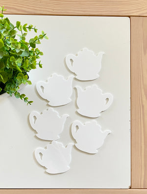 Six white teapot-shaped cutouts arranged on a white surface next to a small green leafy plant on wooden table