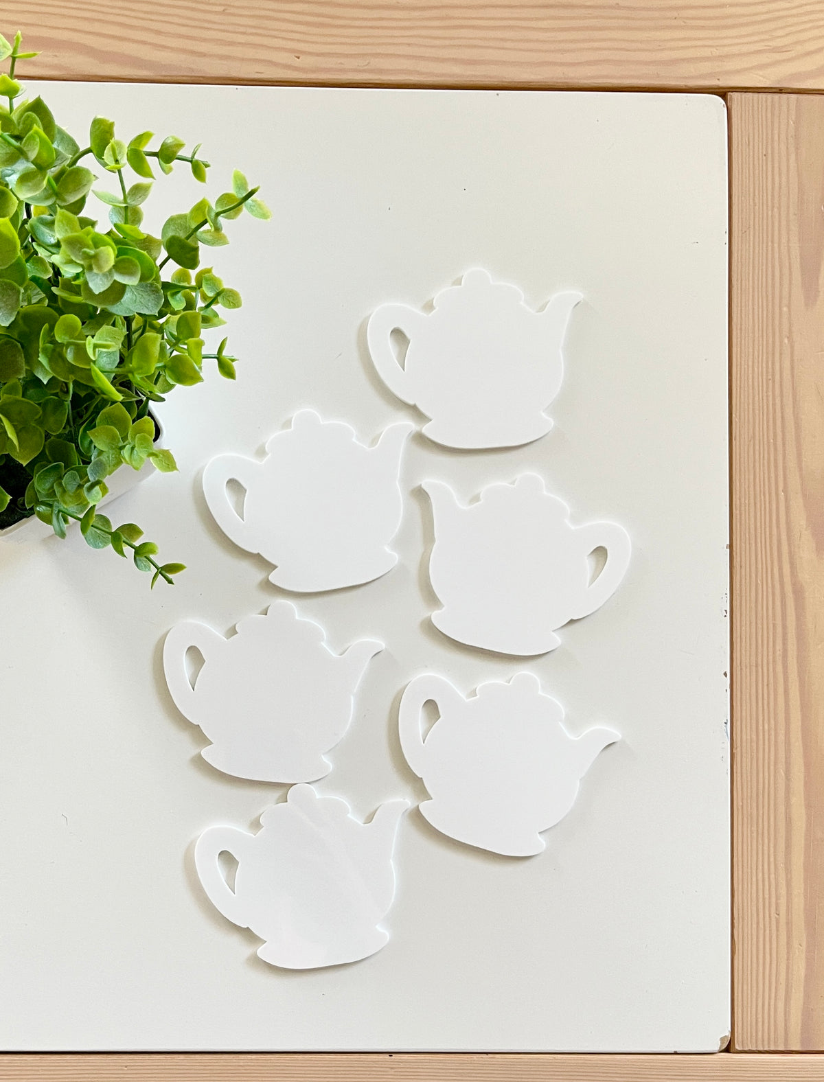 Six white teapot-shaped cutouts arranged on a white surface next to a small green leafy plant on wooden table