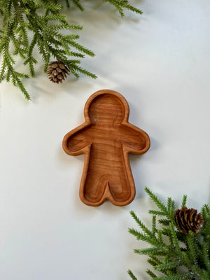 Smooth wooden tray shaped like a gingerbread person surrounded by green pine branches and pine cones on white surface