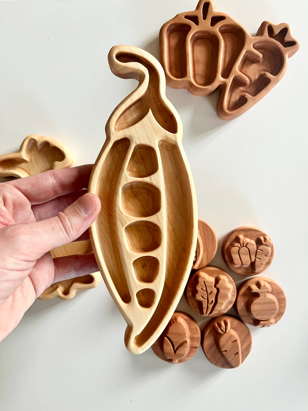 Hand holding wooden tray shaped like a pea pod with other vegetable-shaped trays and wooden tokens on white background
