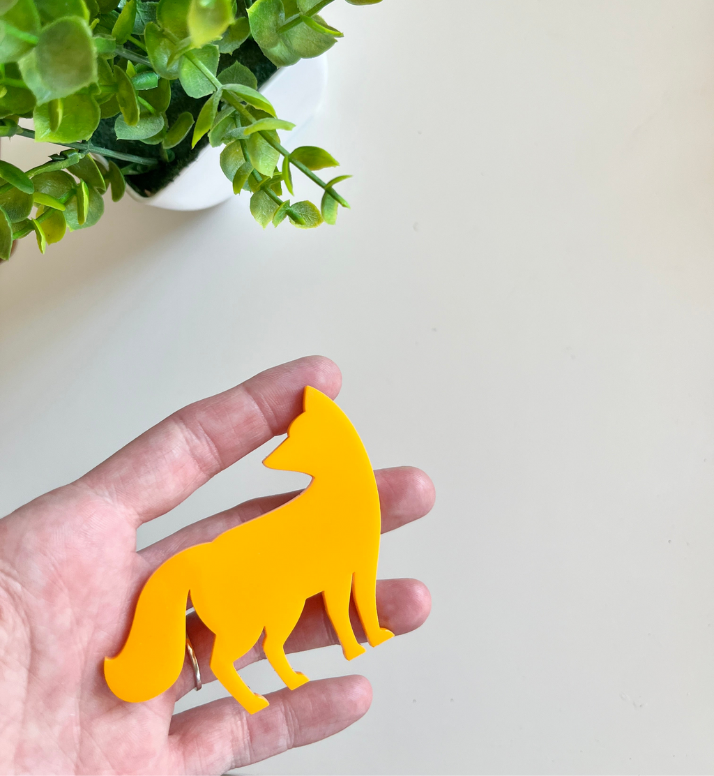 Person holding a smooth bright yellow fox-shaped cutout with green leaves from a potted plant nearby on a light surface