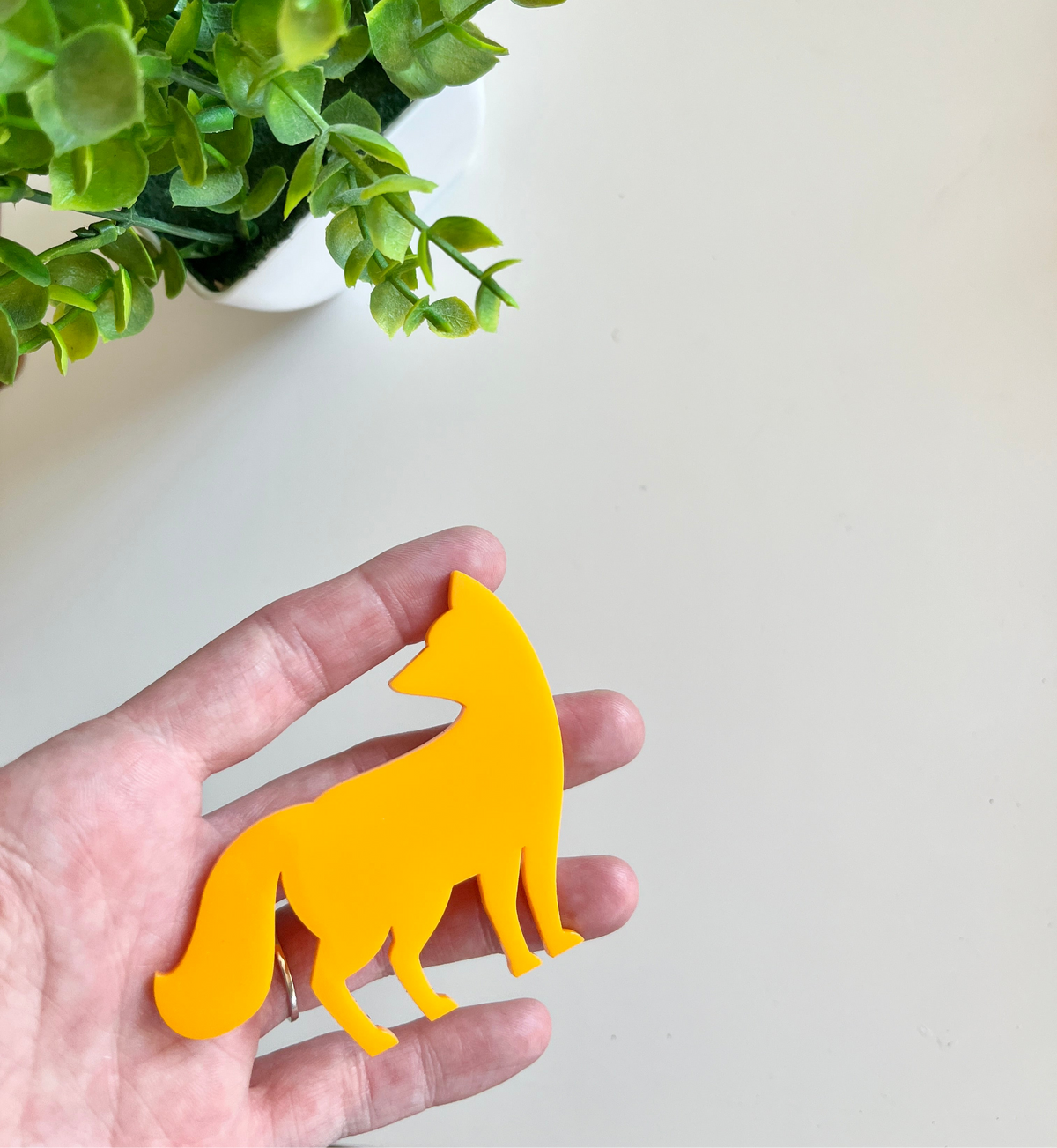 Person holding a smooth bright yellow fox-shaped cutout with green leaves from a potted plant nearby on a light surface